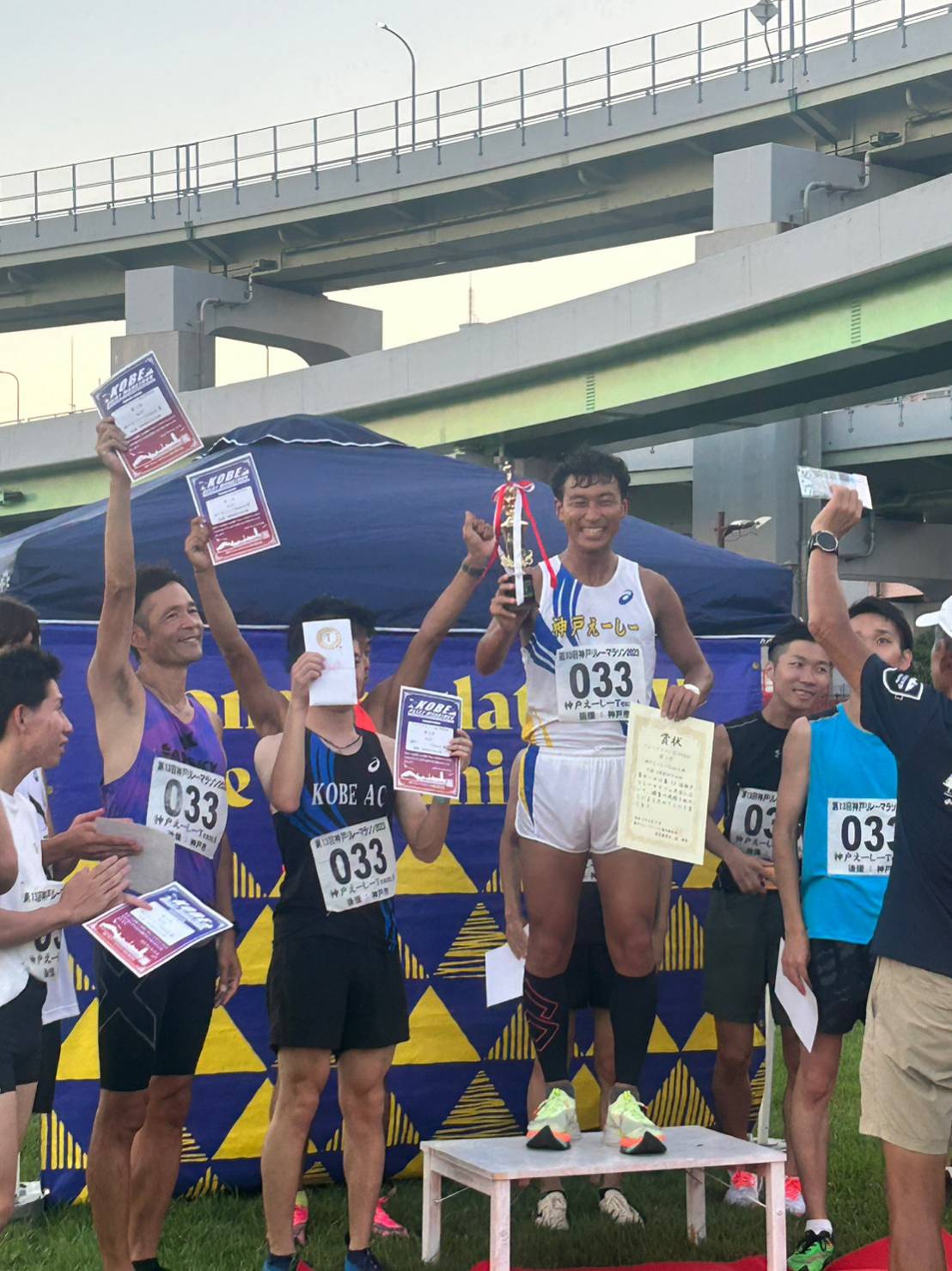表彰台で優勝トロフィーを掲げる神戸えーしーの選手。周囲にメンバーが集まって祝う。