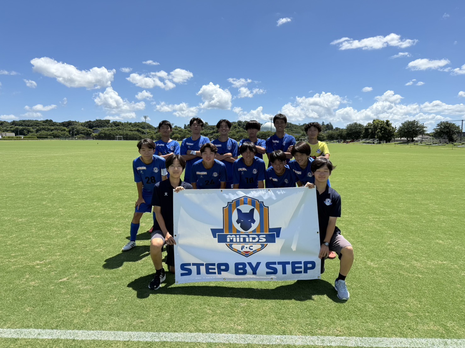 MINDS F.C. の選手たちが STEP BY STEP のチームバナーを掲げて笑顔で並ぶ。