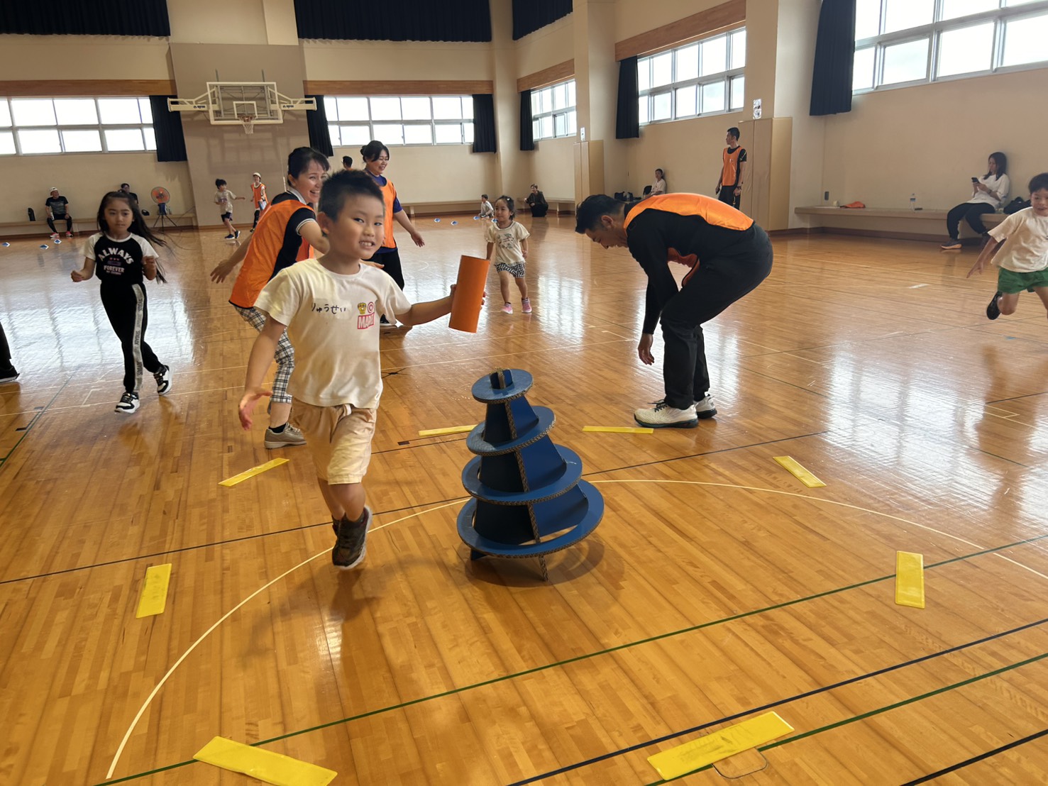 スポーツ鬼ごっこ — 体育館で小さな子どもが青いSpooniゴールに向かって全力ダッシュ、コーチが伴走